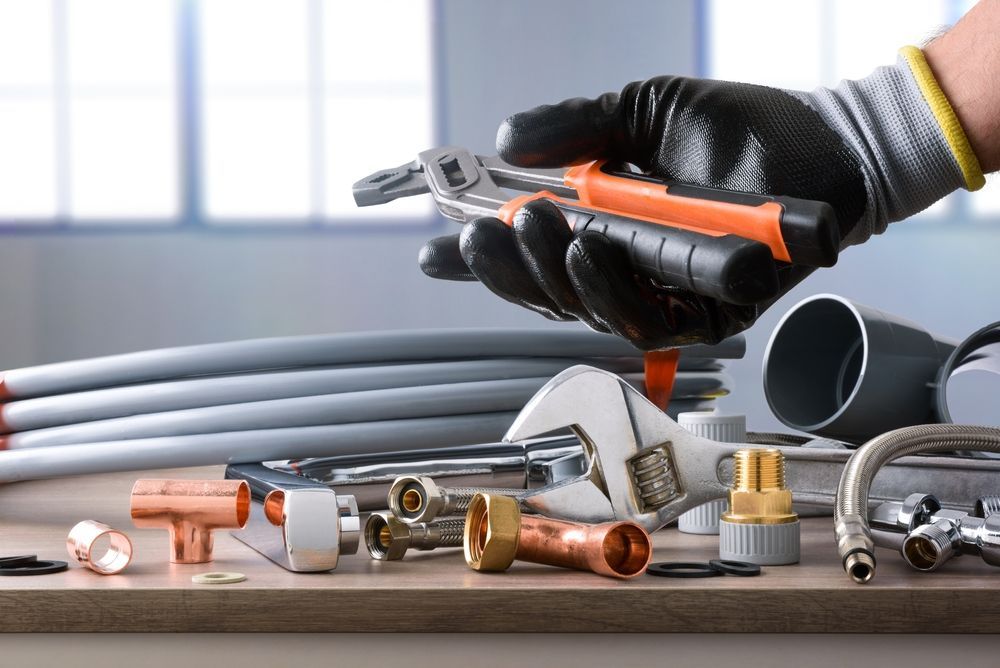 Plumber's hand in black glove holding tools over plumbing supplies on a wood surface.