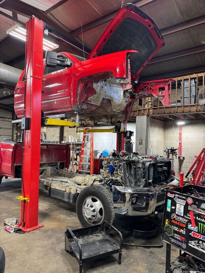 Mechanic working under a raised car hood in a garage, inspecting the engine with tools. | Campbell Automotive