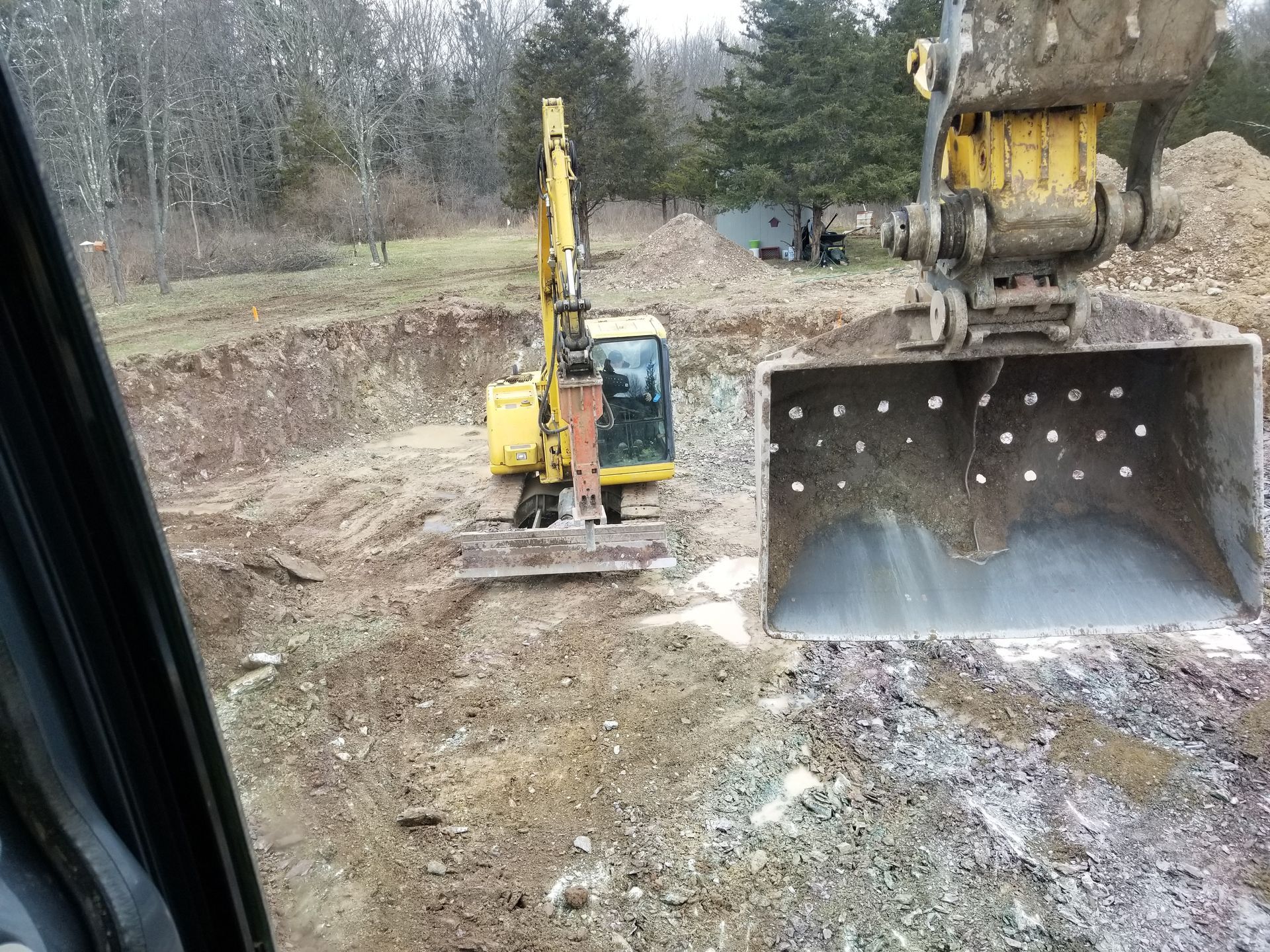A yellow excavator digs in a dirt pit, its bucket visible from the driver's perspective.