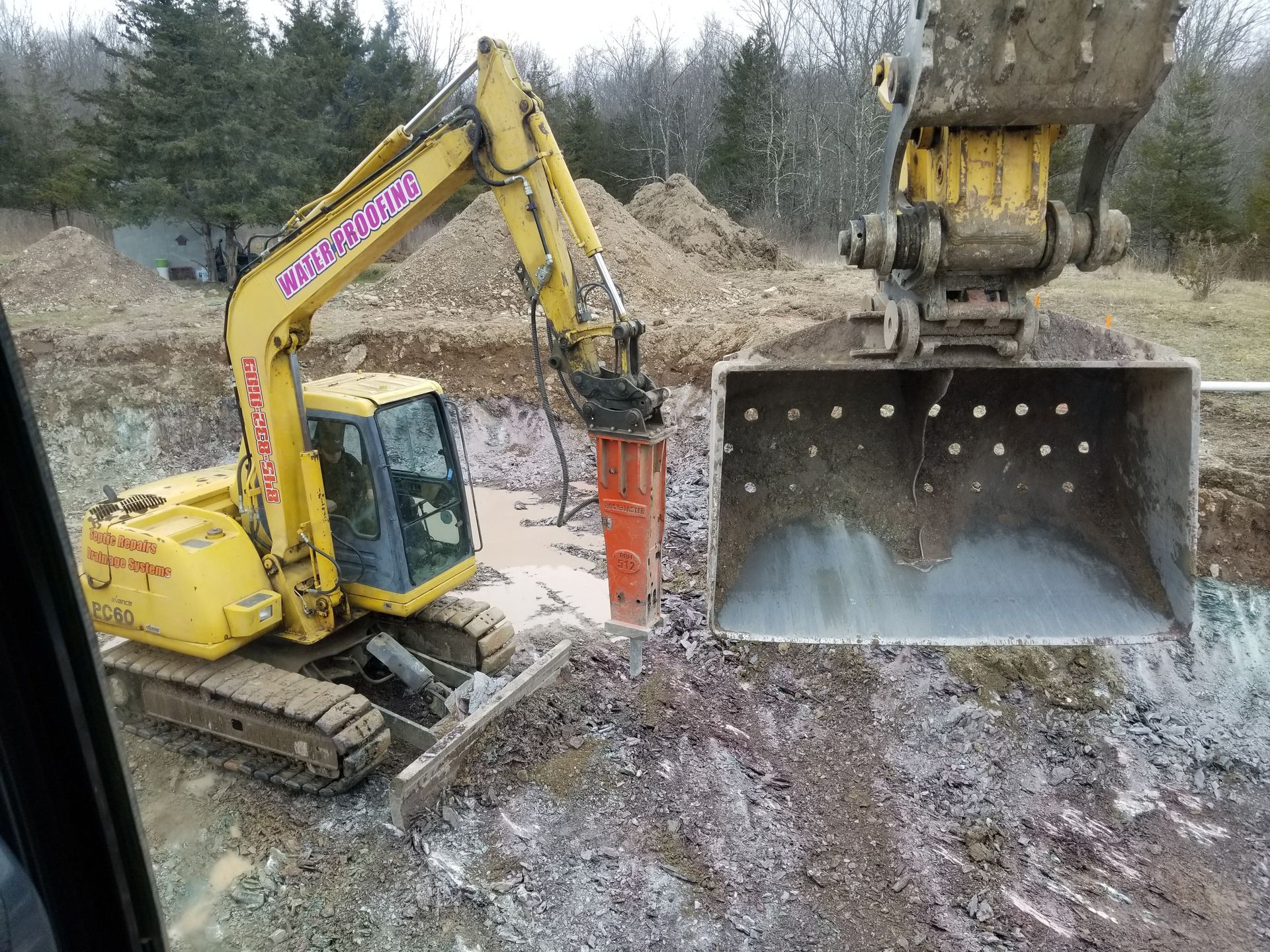 Yellow excavator with jackhammer in a muddy pit, breaking up material.