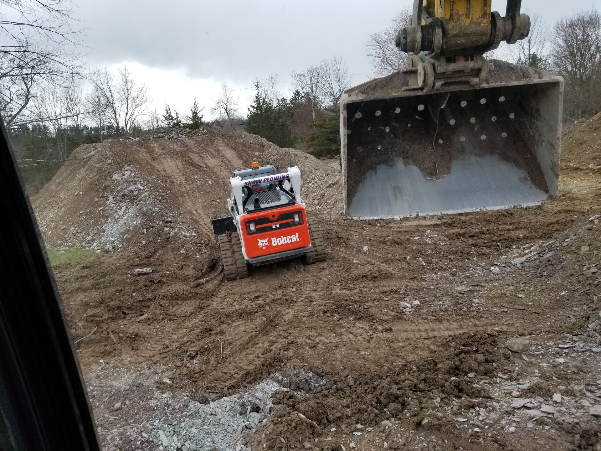 Bobcat skid steer next to an excavator bucket on a dirt pile. Outdoors, overcast.