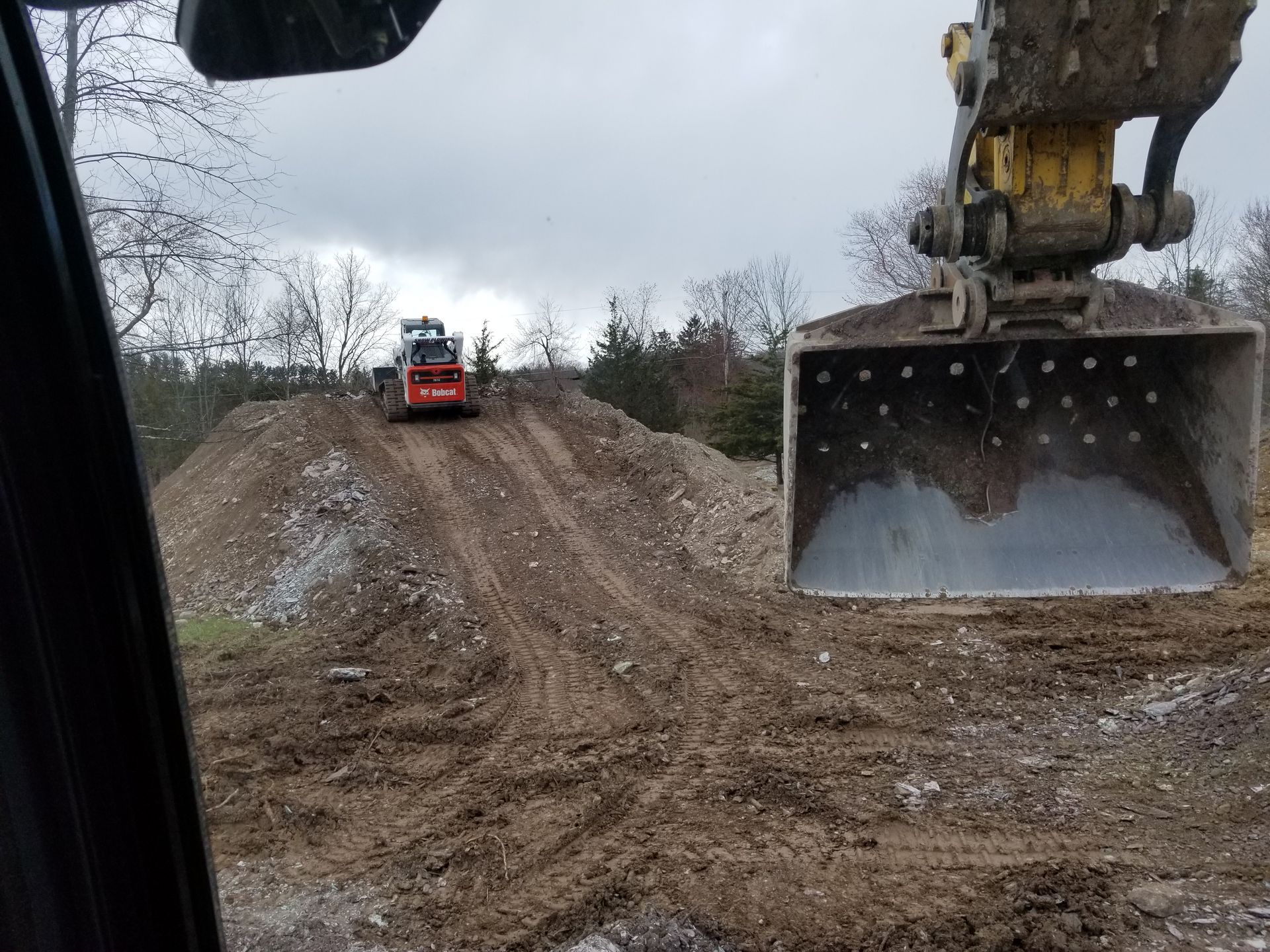 Excavator bucket in foreground, small bulldozer on dirt mound. Cloudy sky, construction site.