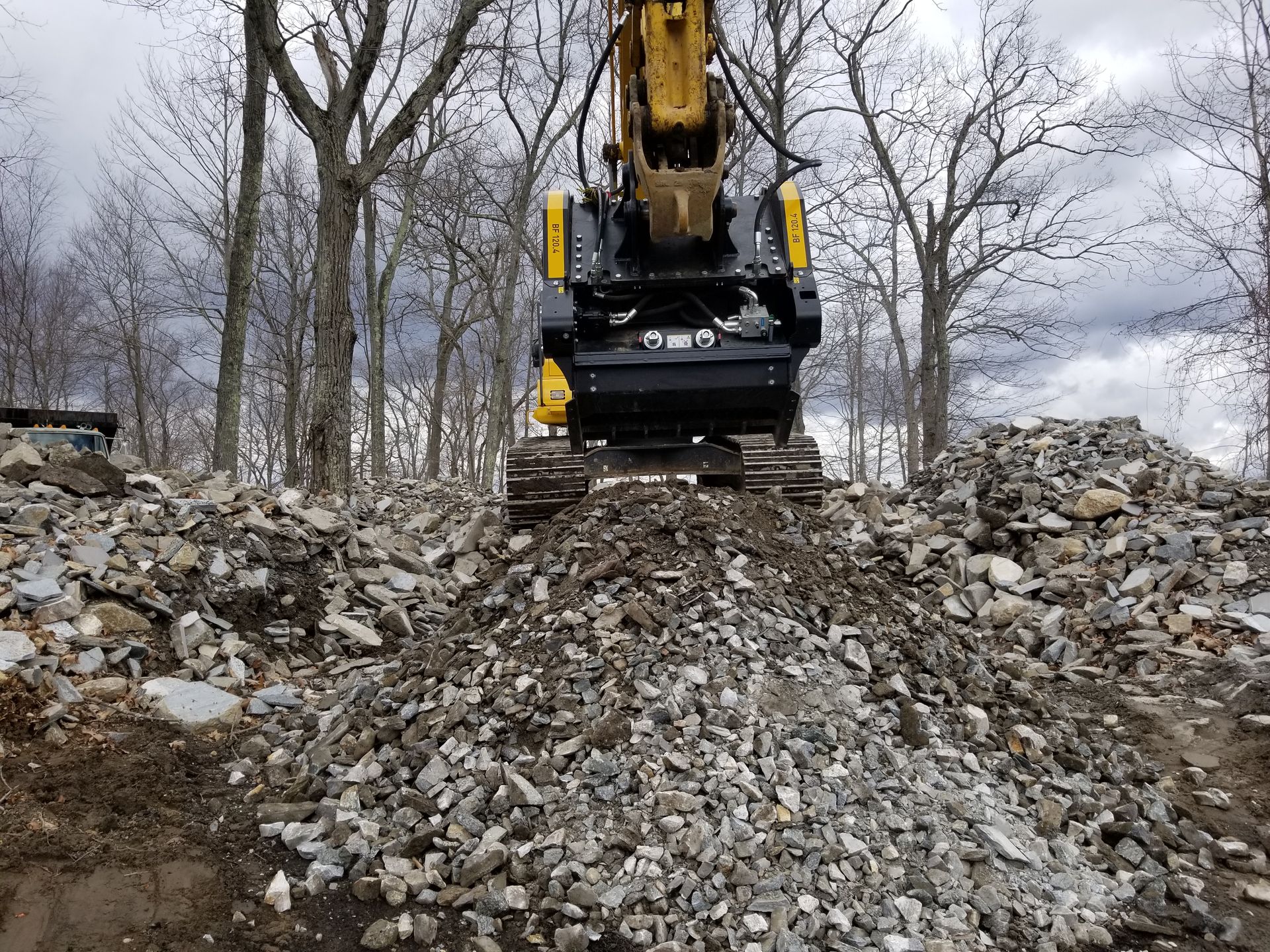 Yellow excavator on pile of rubble in a wooded area, cloudy sky.