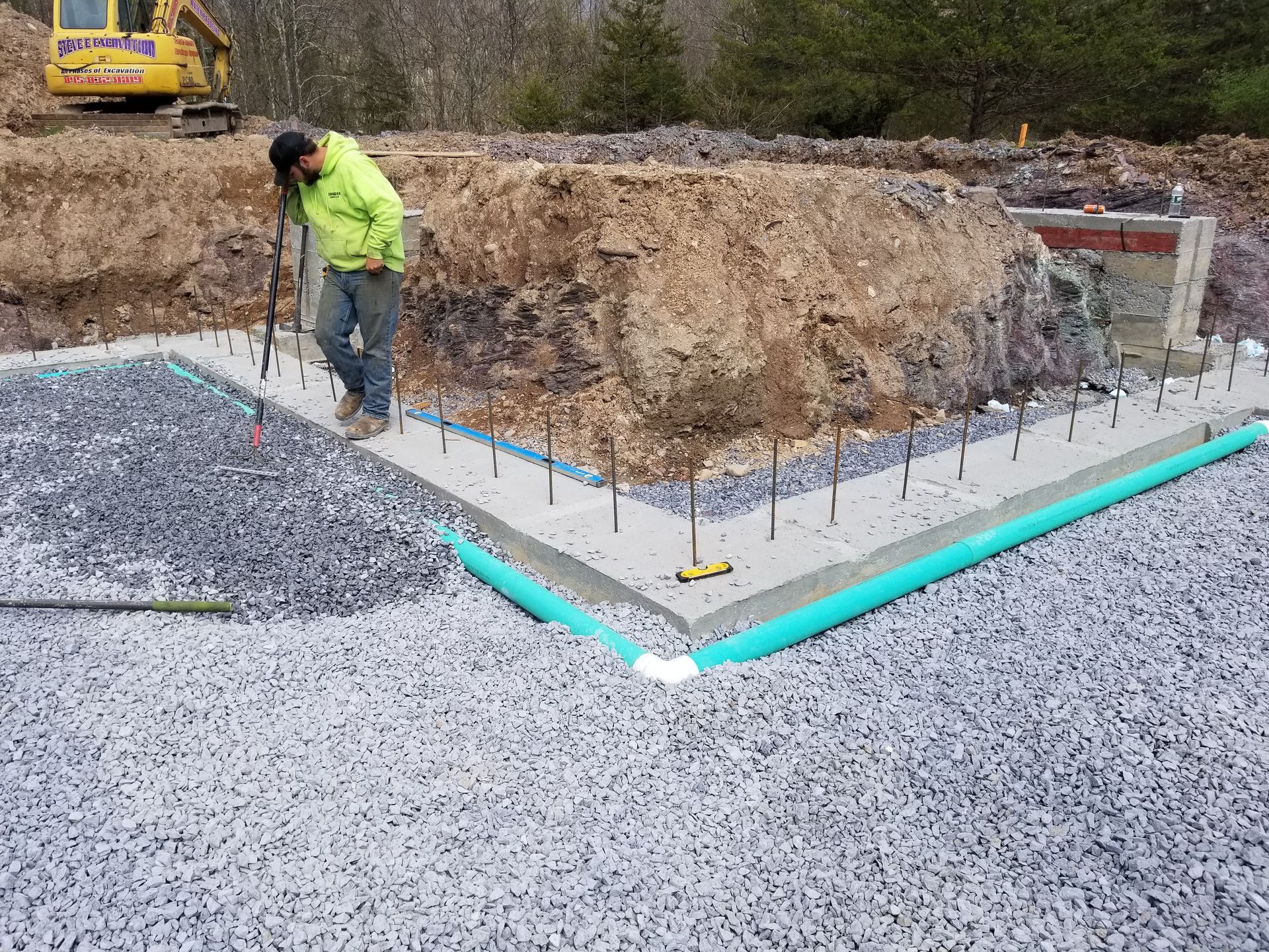 Construction worker checking a concrete foundation corner. Blue PVC pipe, rebar, gravel, and an excavator are visible.