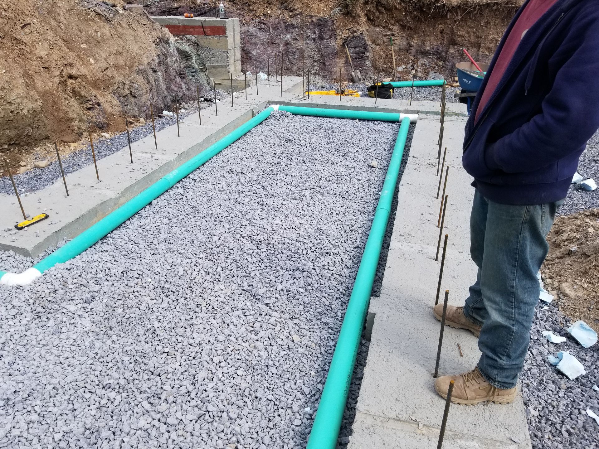 Construction site with a rectangular gravel bed bordered by concrete and green pipes; a person stands nearby.
