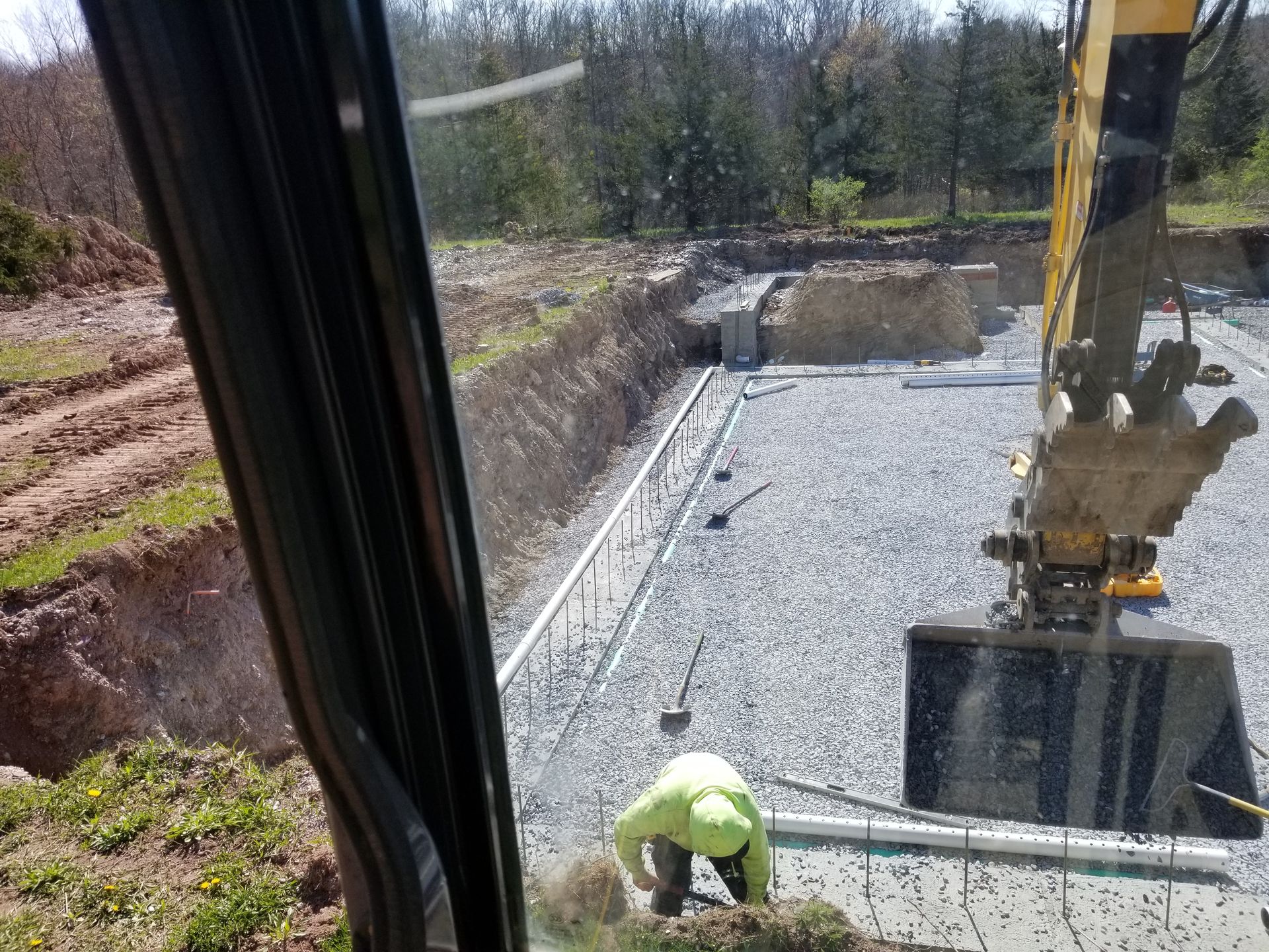Construction site: An excavator, worker, and foundation preparations with gravel and pipes.