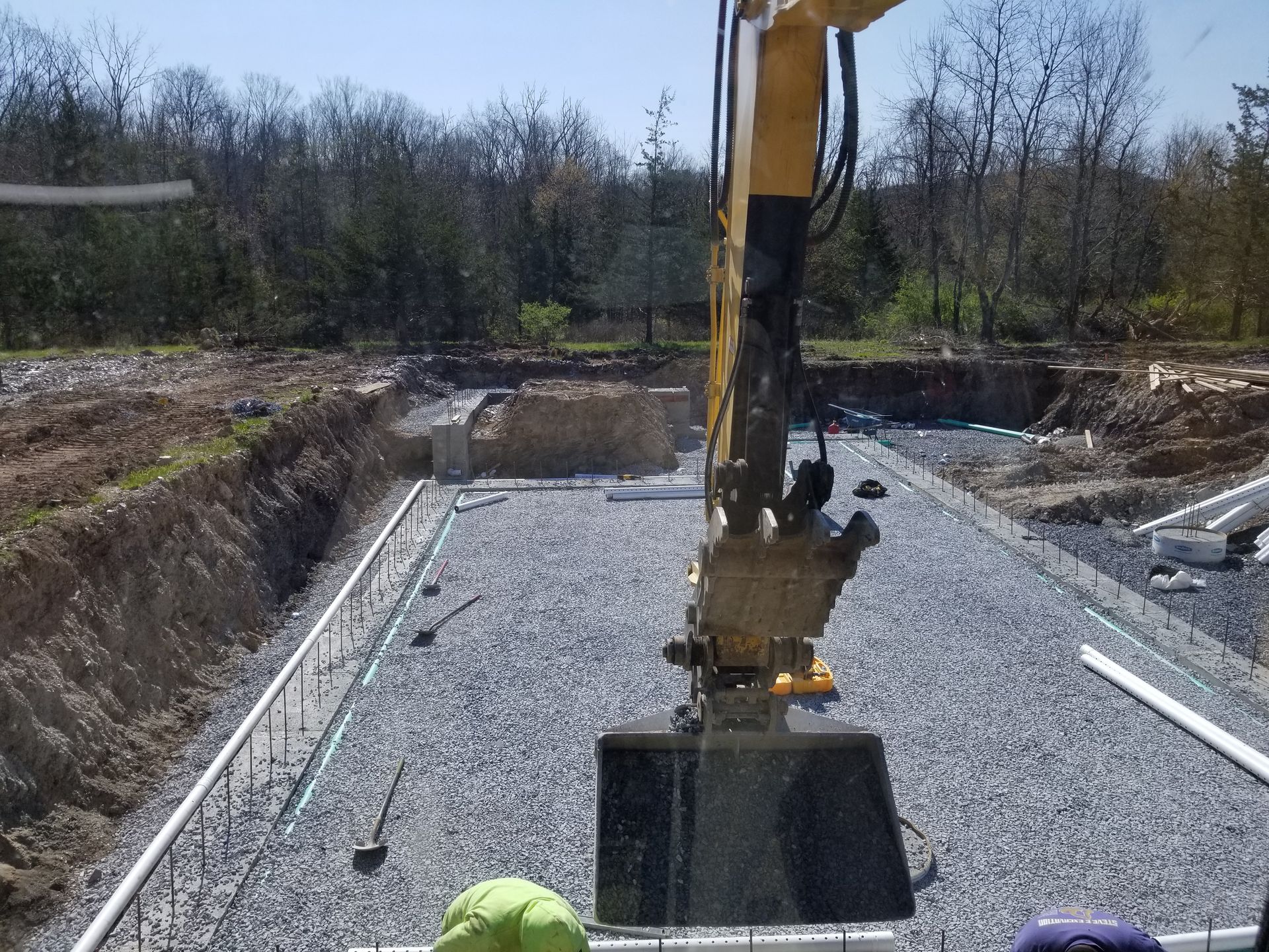 Excavator over a gravel-covered construction site, with PVC pipes and foundation forms visible.