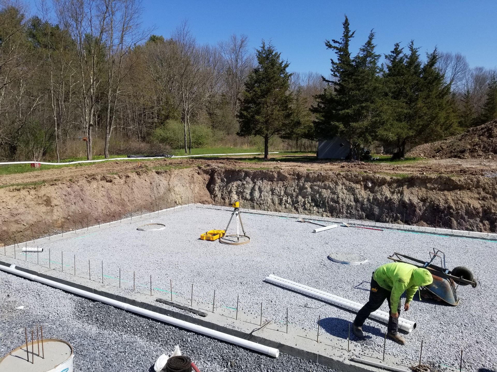 Construction site: workers install pipes on a gravel bed within a foundation pit, sunny day.