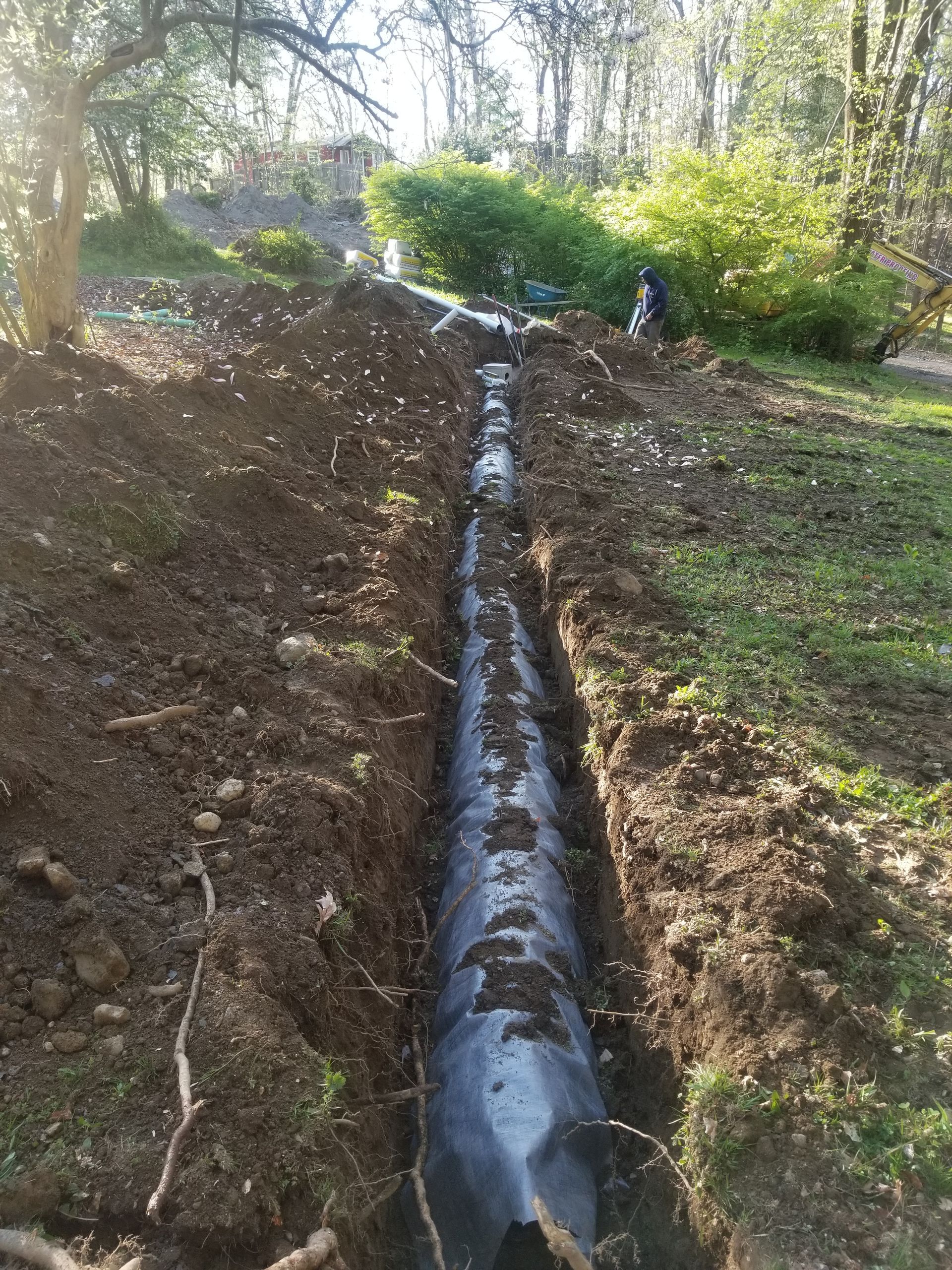 A long trench with black pipe laid in it, dug in brown soil, in a grassy area.