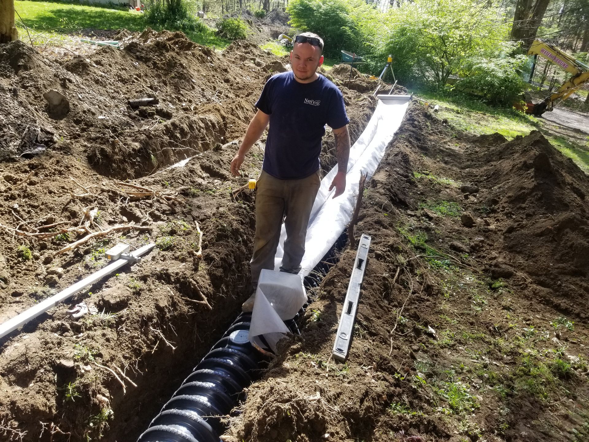 Man installing drainage pipe in a trench. Black pipe, white fabric, level, dirt, and greenery.