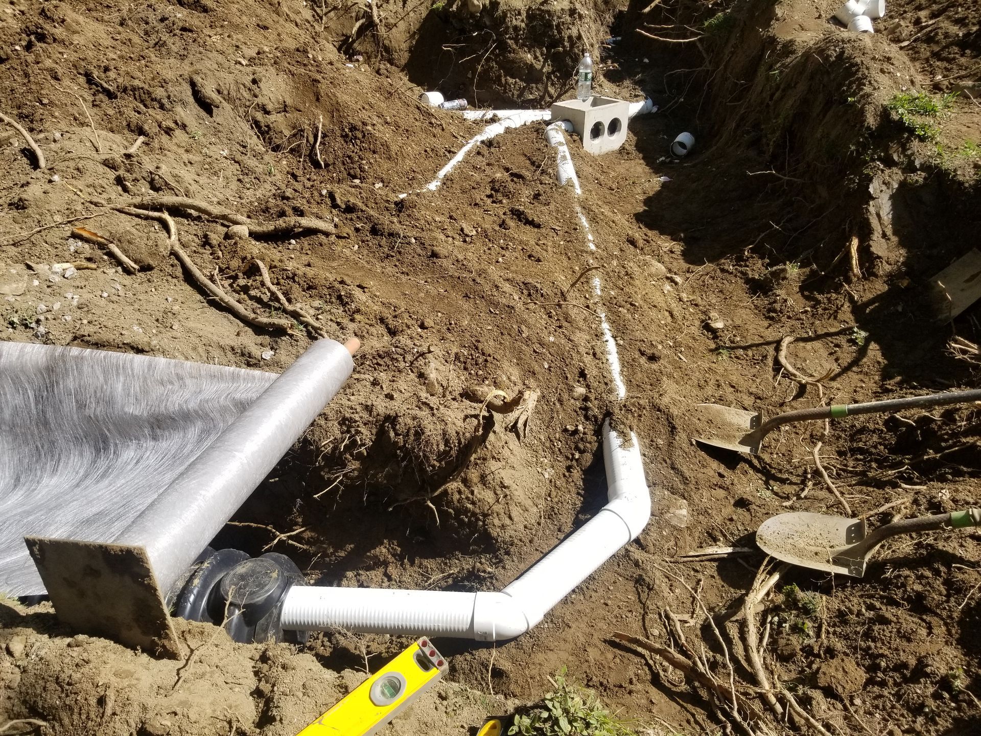Pipes and a drain system in a dug-out area, with a level and tools, likely for landscaping or drainage work.