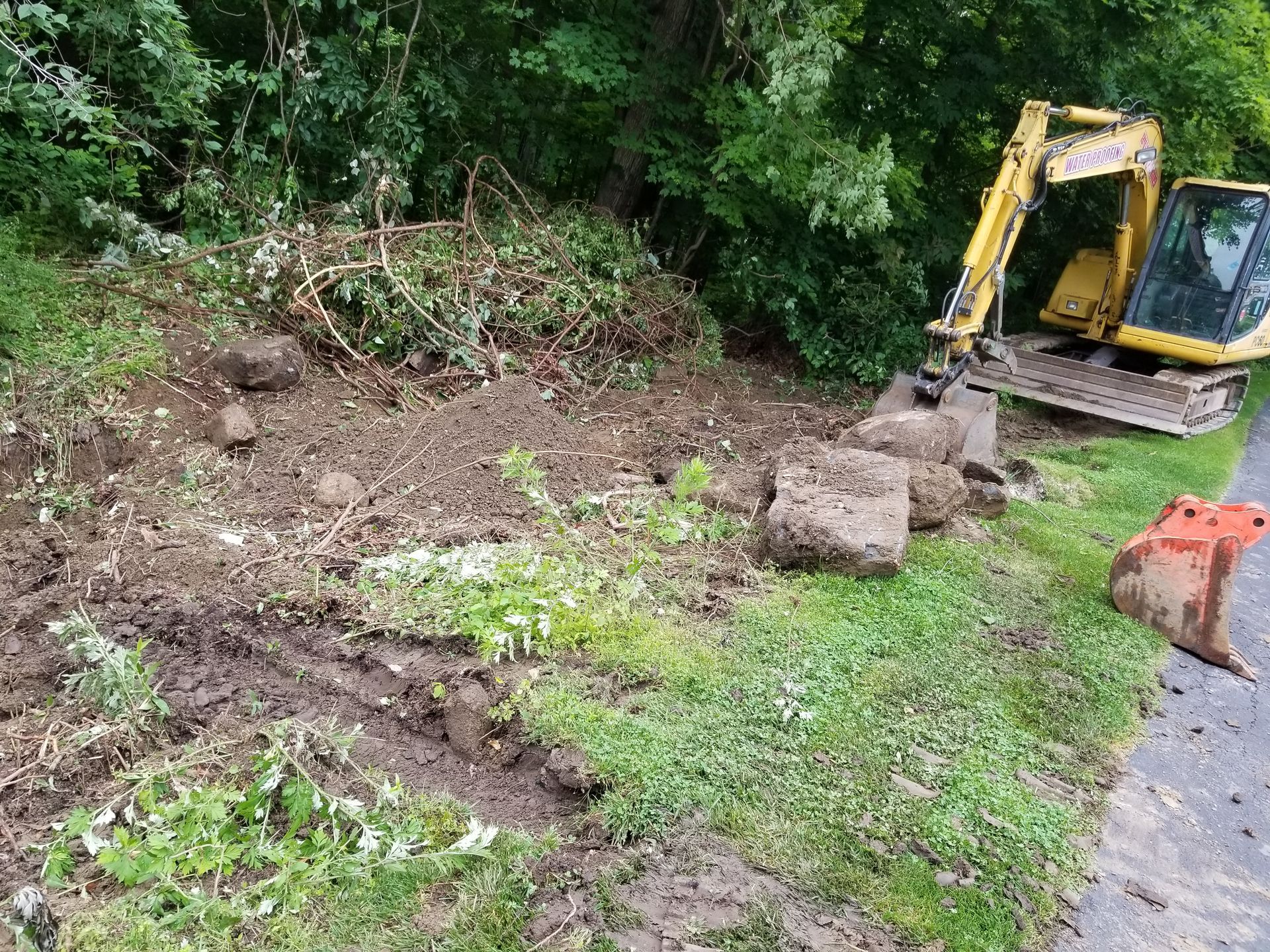 Yellow excavator next to debris pile near a road, grass, and trees.