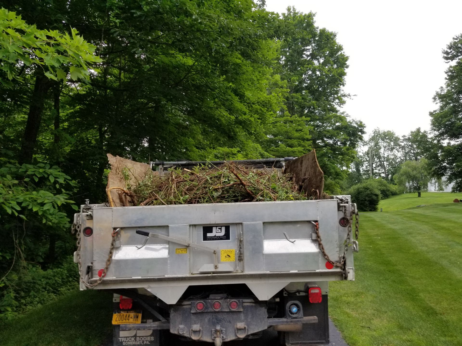 A silver dump truck bed filled with yard waste, parked on a grassy area, surrounded by trees.