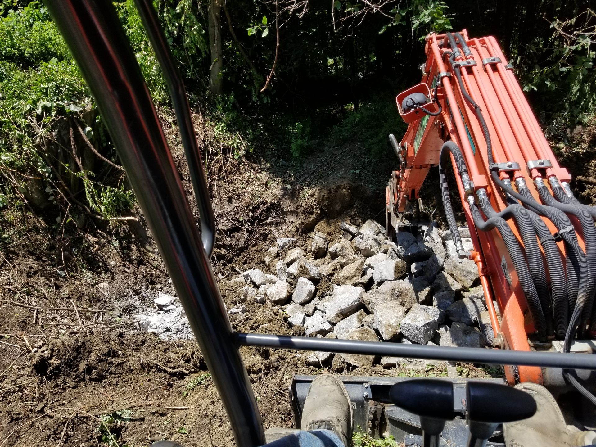 View from excavator cab as it moves rocks on a hillside path. Orange machinery contrasts with green foliage.