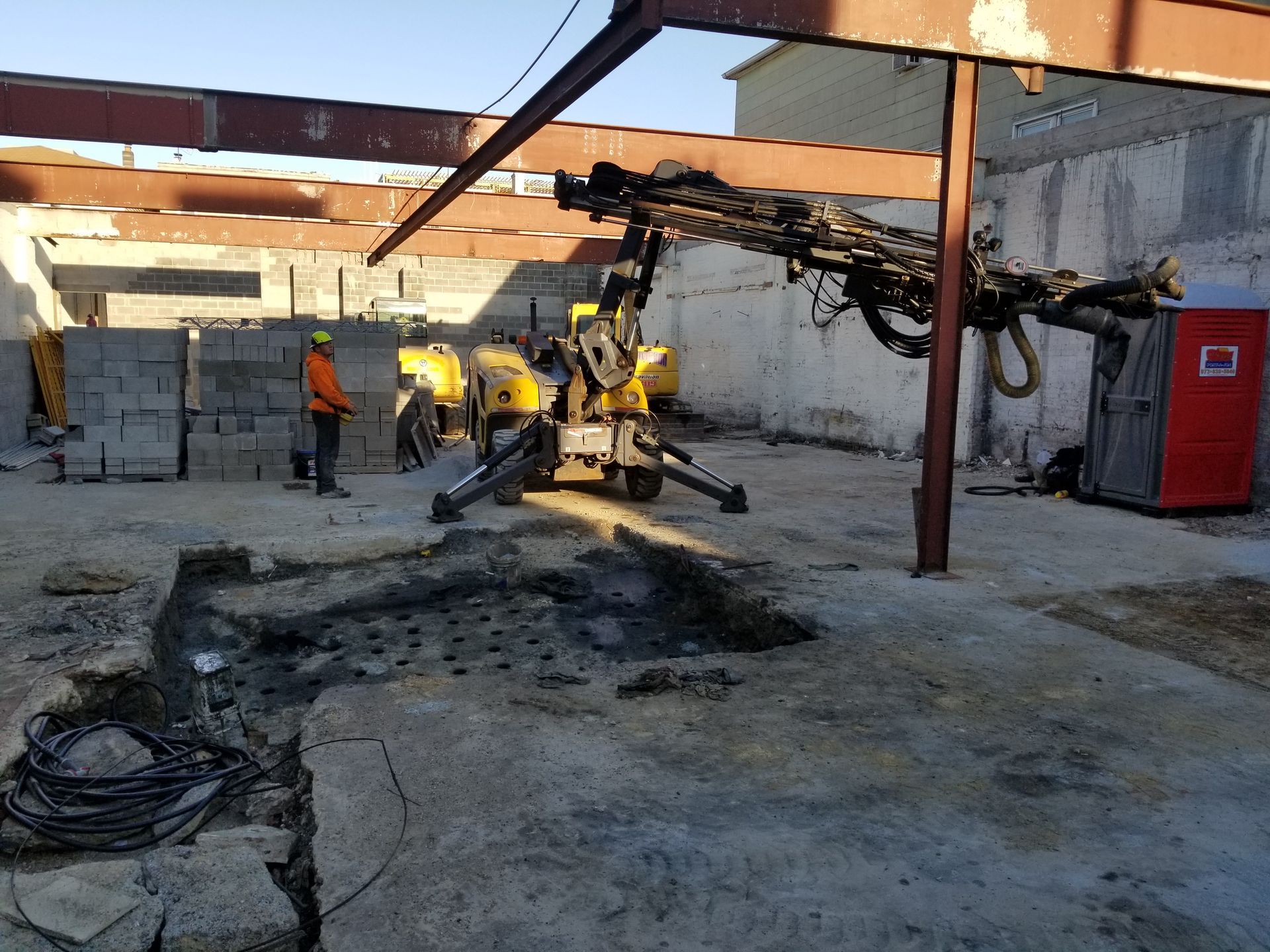 Construction site with drilling equipment and worker. Concrete floor, yellow machinery, and exposed beams.