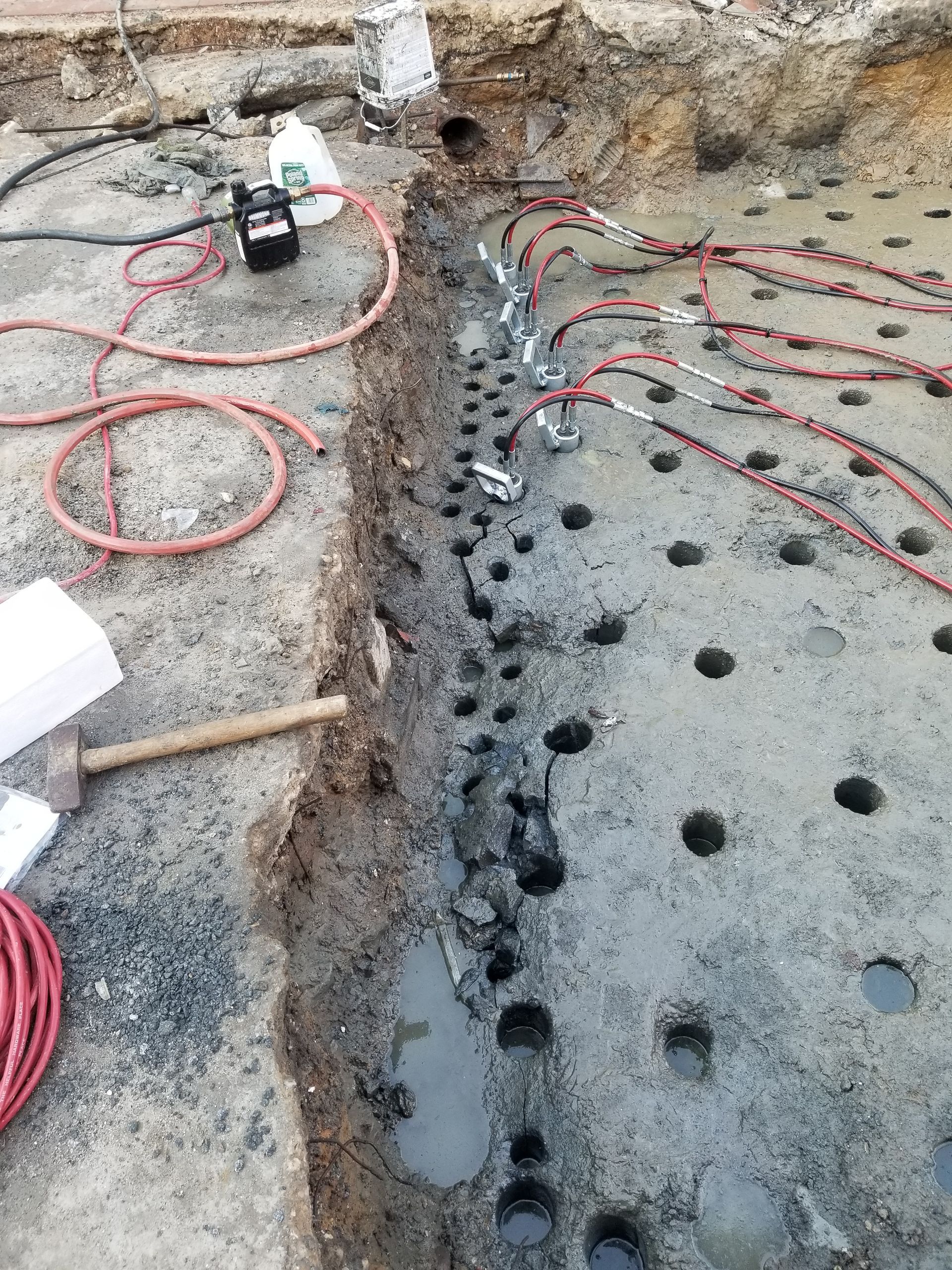 Dug trench with many drilled holes; red and black wires attached to the holes, tools, and equipment on the ground.