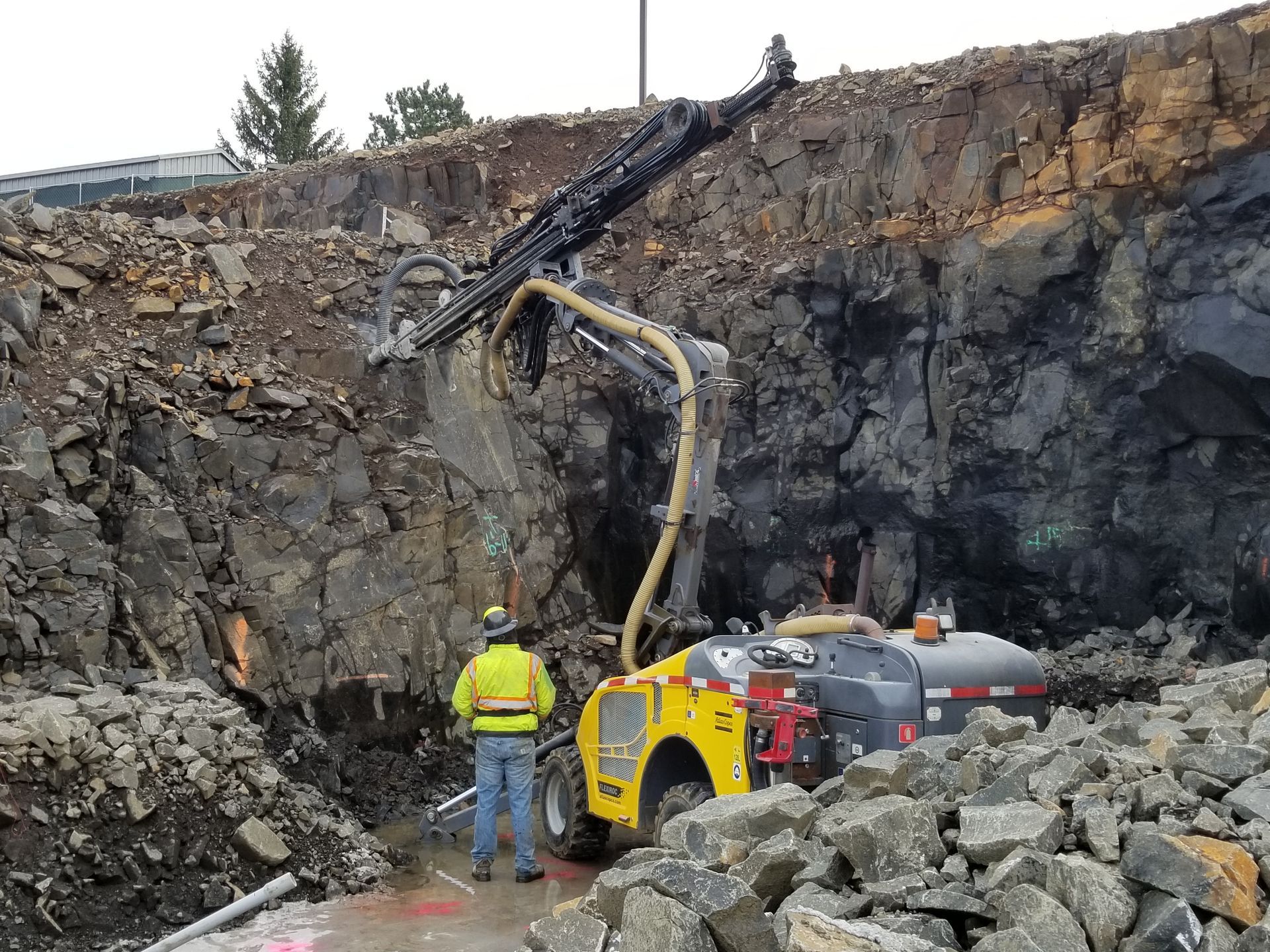 A worker in yellow vest operates a drill on a rocky construction site.