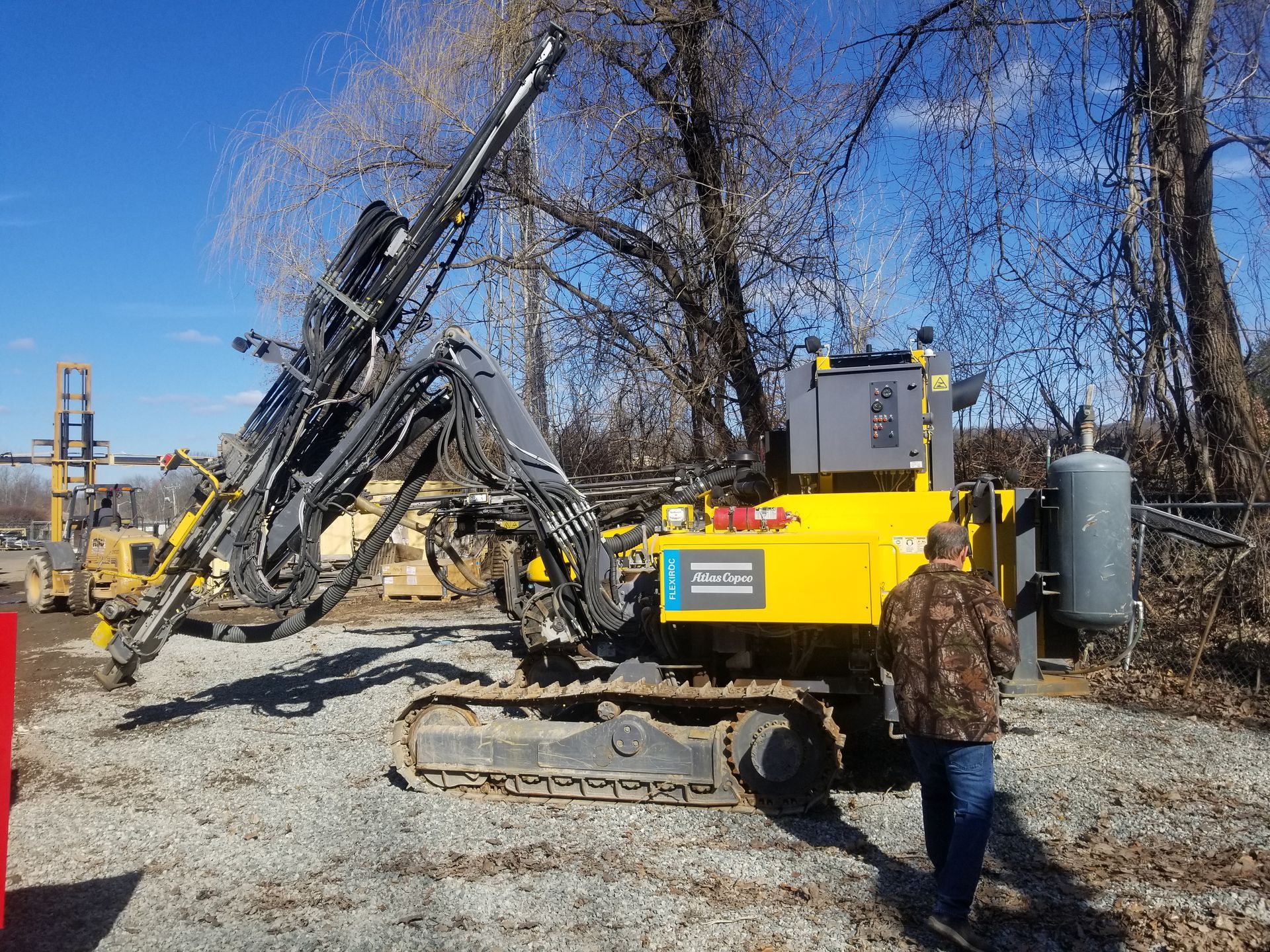 Yellow and black drill rig with a person in camo jacket, outdoors on gravel.