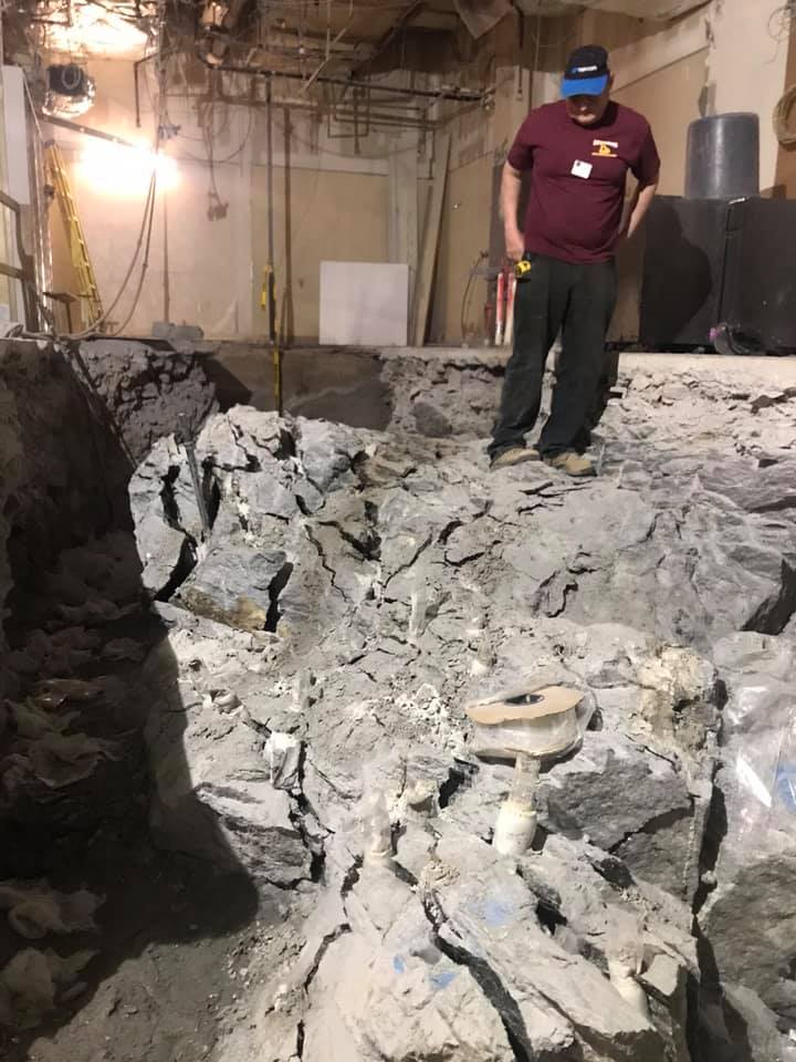 Man standing near a large, rough hole in a concrete floor. Interior, construction site.