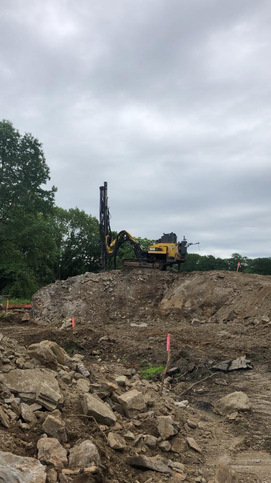 Construction site with yellow drilling machine atop a mound of dirt and rocks under a cloudy sky.