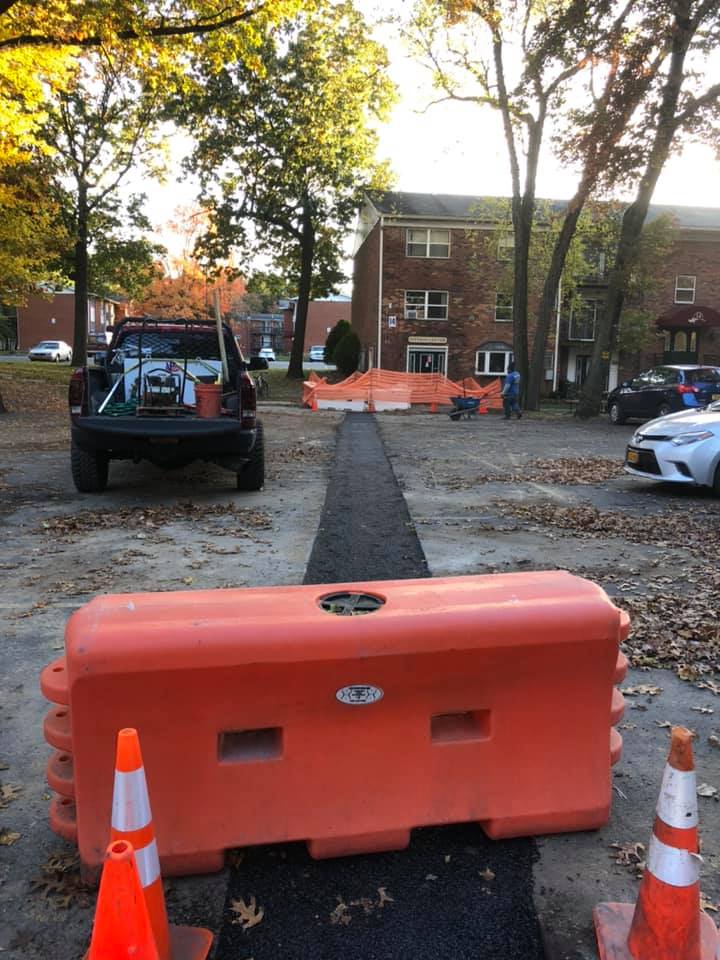 Orange barriers and cones block a newly paved path in front of an apartment building, with a work truck in the background.