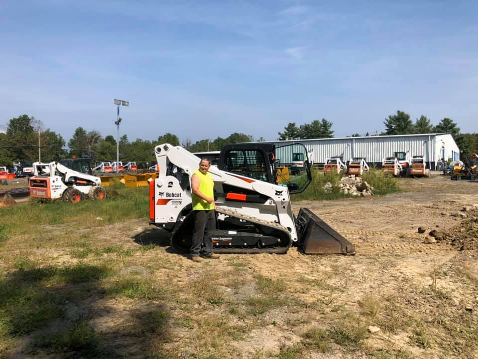 Man in safety vest stands by a Bobcat compact track loader on a dirt lot with other construction equipment.