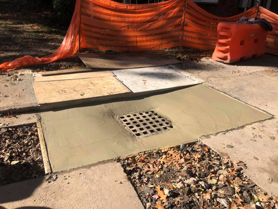 Concrete work around a sewer grate, with orange safety fencing, wooden and cardboard panels on sidewalk.