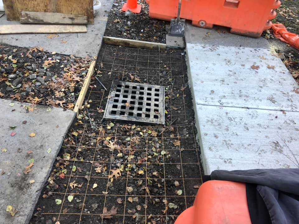Sidewalk under construction; grate sits in the ground surrounded by metal grid, orange barriers in view.