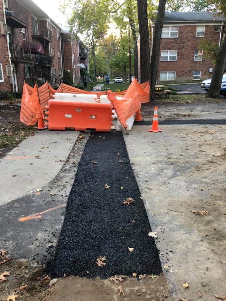 Newly paved pathway blocked by orange construction barriers, near apartment buildings.
