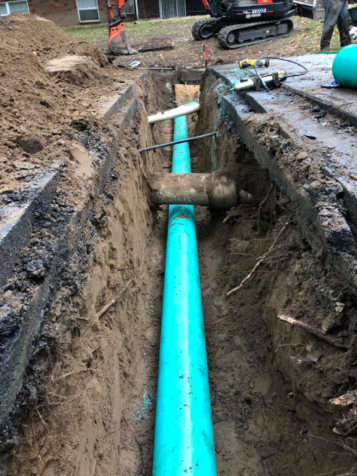 A trench with blue PVC pipe being installed next to asphalt. An excavator is in the background.