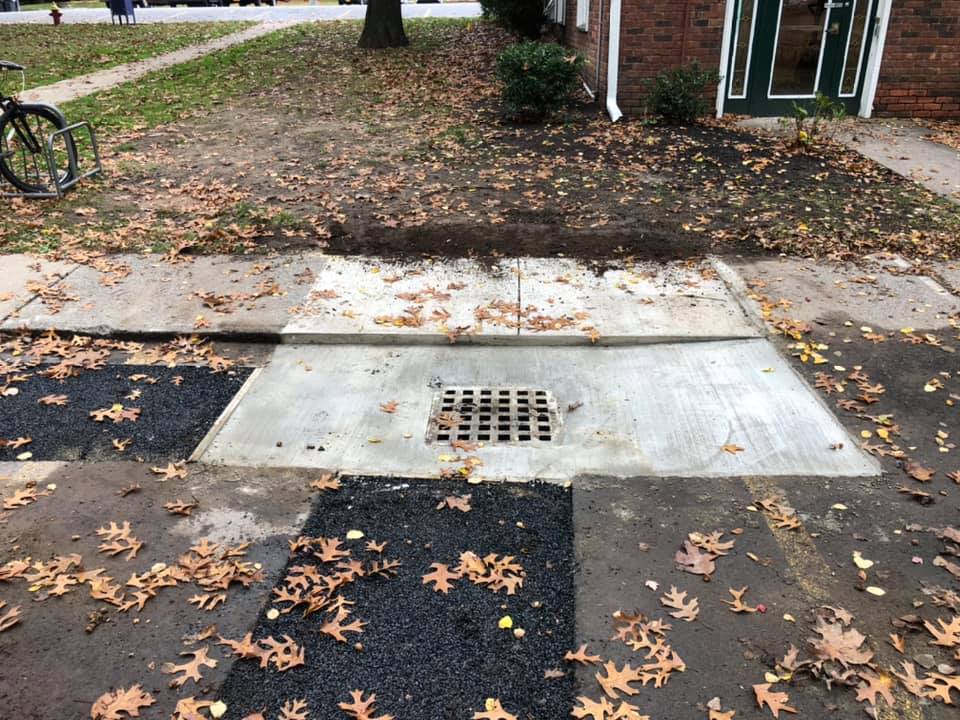 Concrete drainage area with metal grate in sidewalk, surrounded by asphalt and leaves. Bicycle in background.