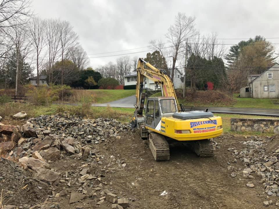 Yellow excavator on a dirt path next to a pile of rocks. Houses and trees in the background under an overcast sky.