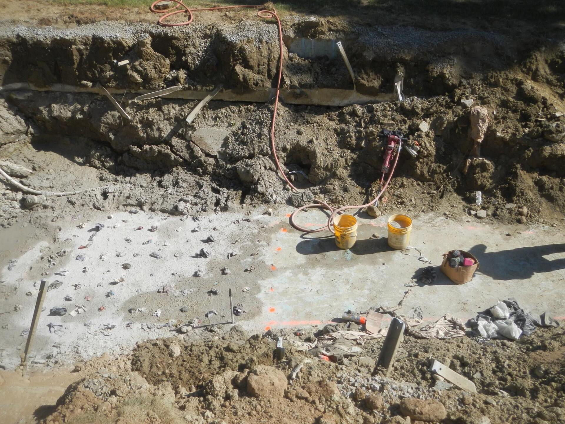Construction site: trench with concrete base, dirt walls, buckets, tools, and wires.