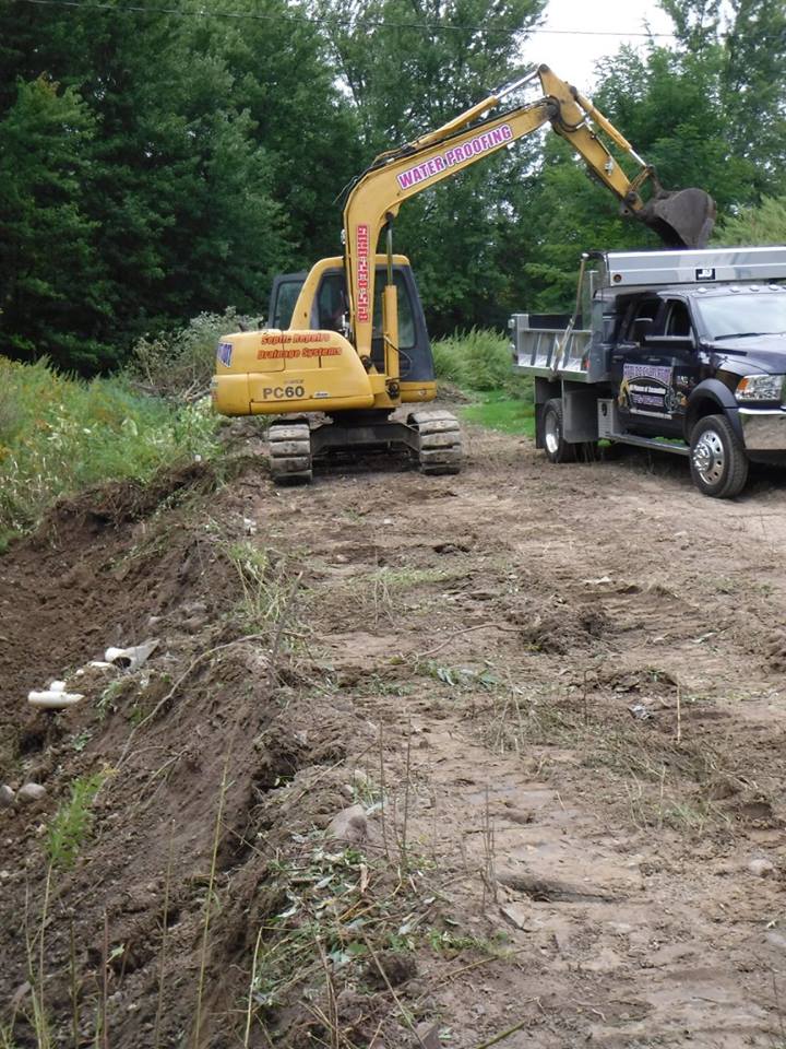 Yellow excavator loading dirt into a black truck on a dirt road, trees in the background.