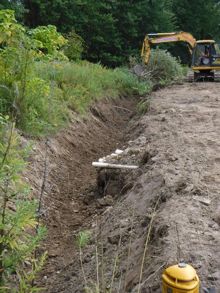 Long trench in dirt, with a backhoe in the background, likely for laying pipe. Green trees and grass border.