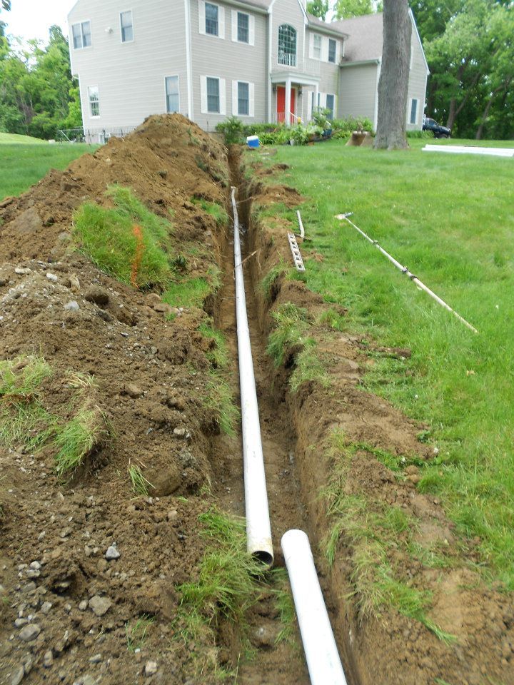 Trench dug in a yard with white PVC pipes laid inside, house in background.