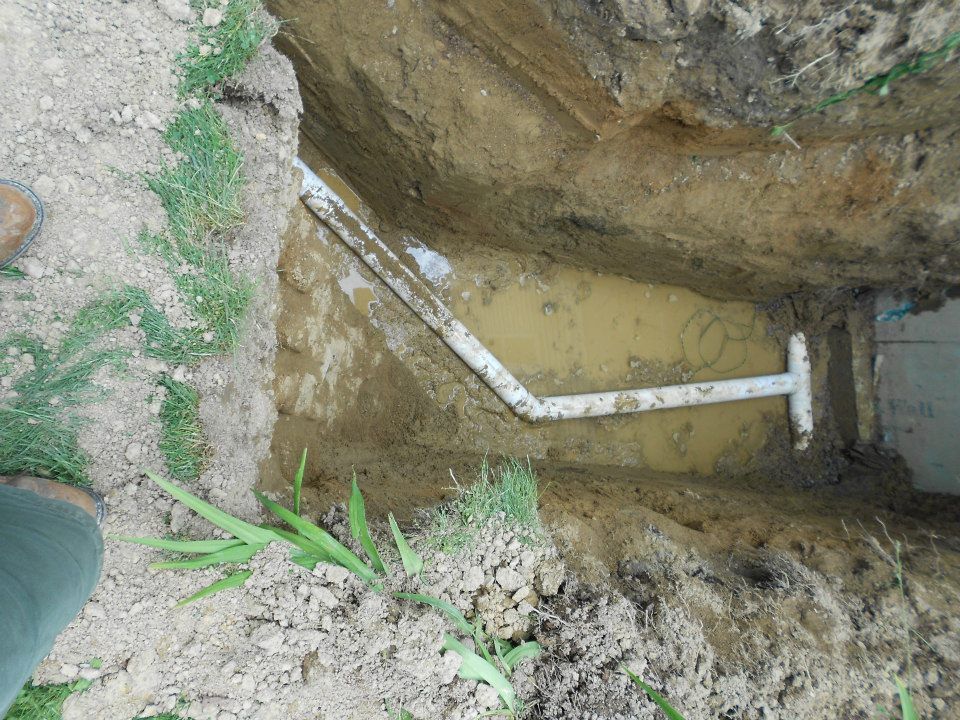 A trench dug in the dirt with PVC pipes running through it, alongside a concrete structure.
