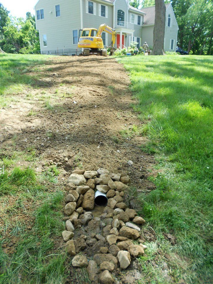 A drainage system is being installed in a grassy yard. A yellow excavator sits by a house.