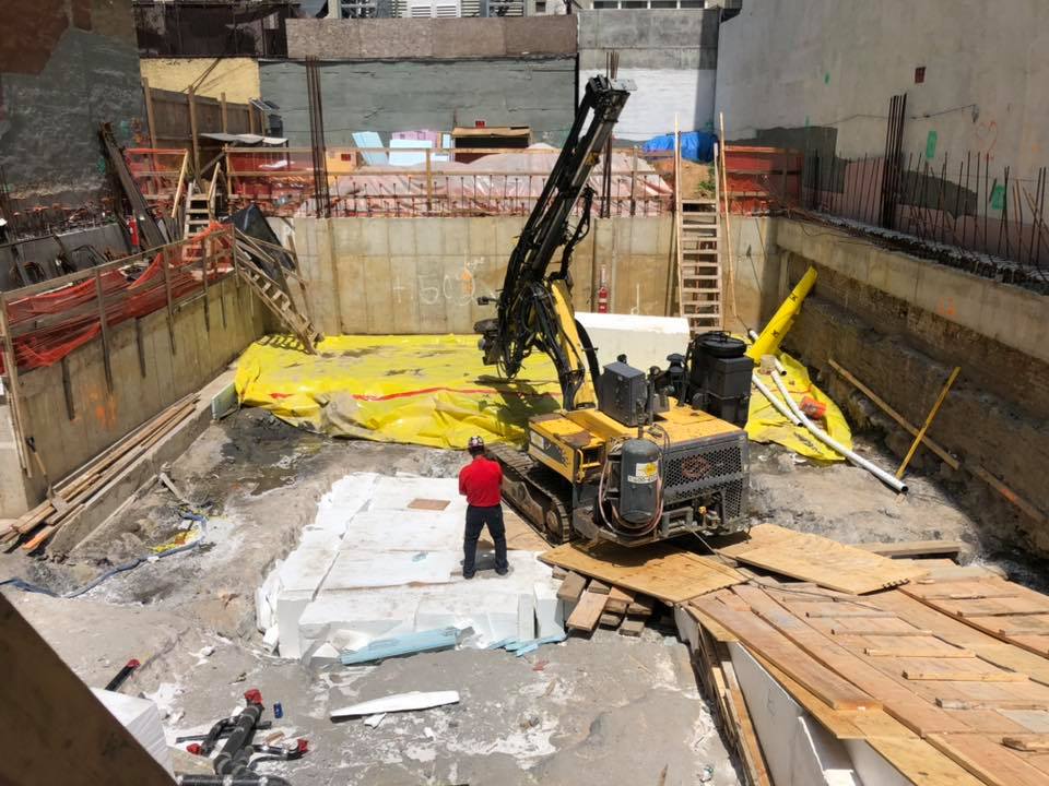 Construction site with drilling equipment; worker in red shirt observes. Concrete walls and ground.