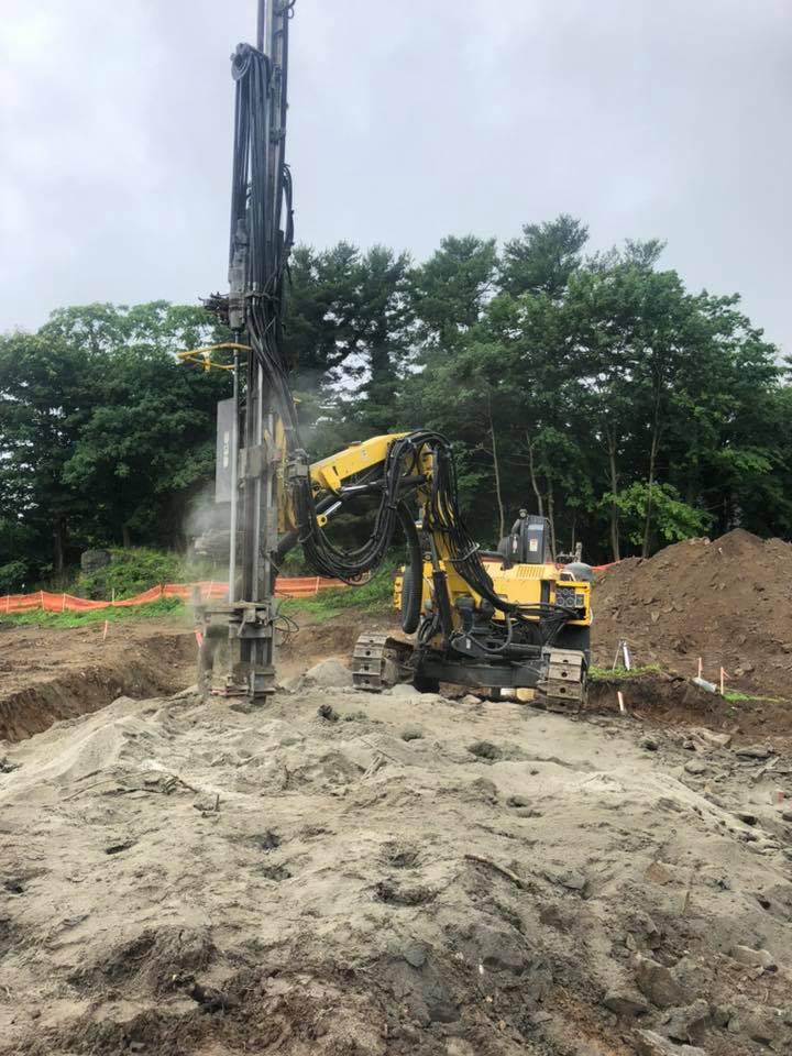 Yellow drilling machine on construction site, operating. Grey ground with dirt pile, green trees in background.