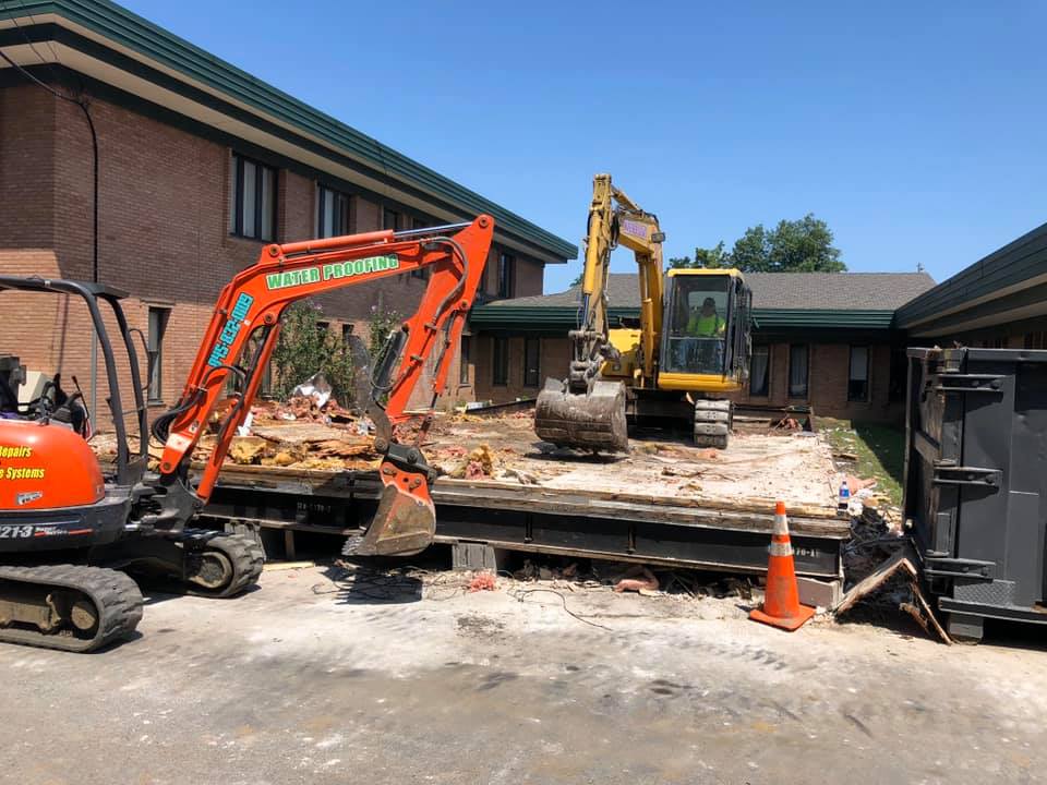 Two excavators demolishing a structure next to a brick building on a sunny day.
