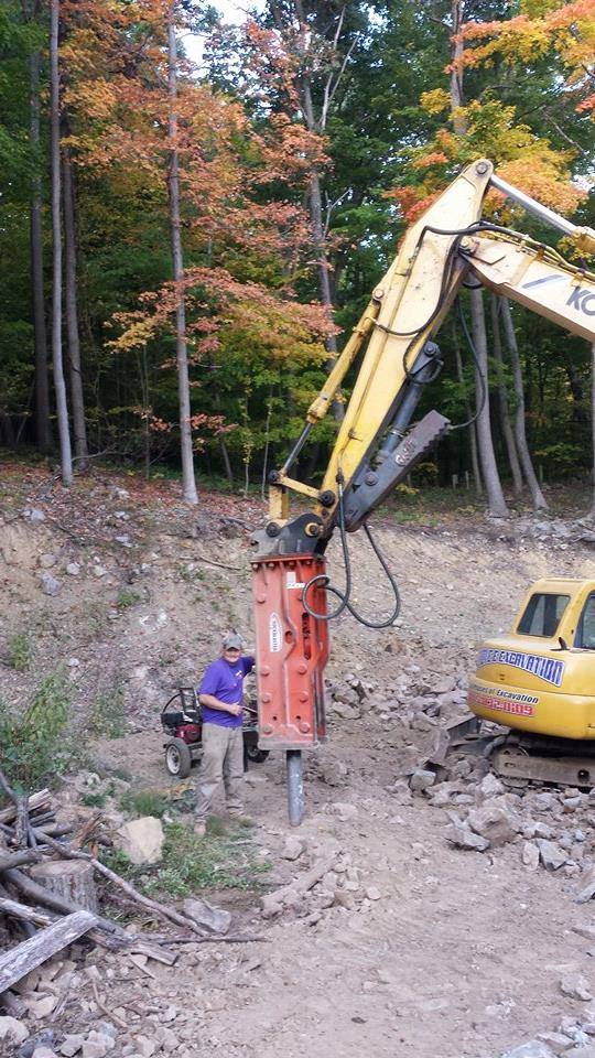 Man operating excavator with jackhammer attachment outdoors on a rocky hillside.