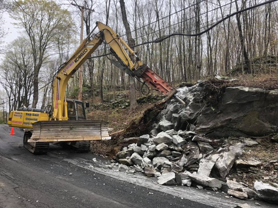 Yellow excavator breaking rocks on a roadside with a wooded backdrop.
