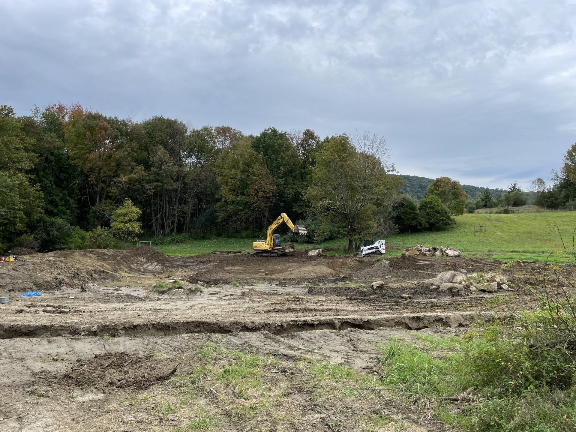 An excavator and a skid steer clearing dirt in a field, trees in the background, under a cloudy sky.
