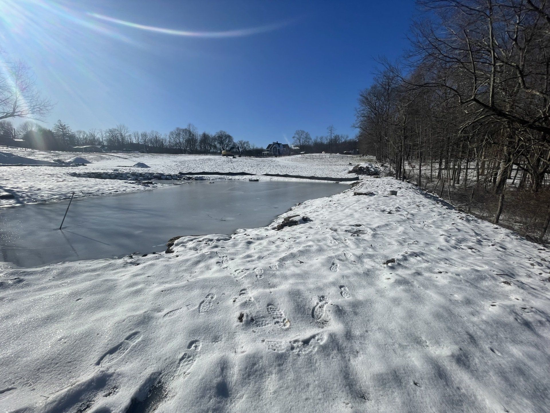 Snow-covered landscape with a frozen pond under a bright blue sky and bare trees.
