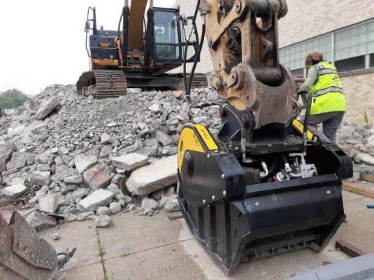 Excavator with demolition attachment sorting debris pile; worker in vest.