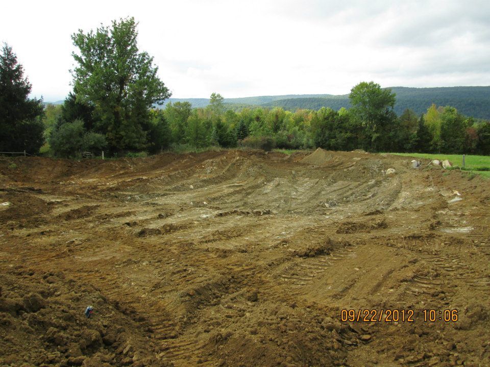 Dirt landscape, recently excavated, with tire tracks. Trees and hills in the background under a cloudy sky.