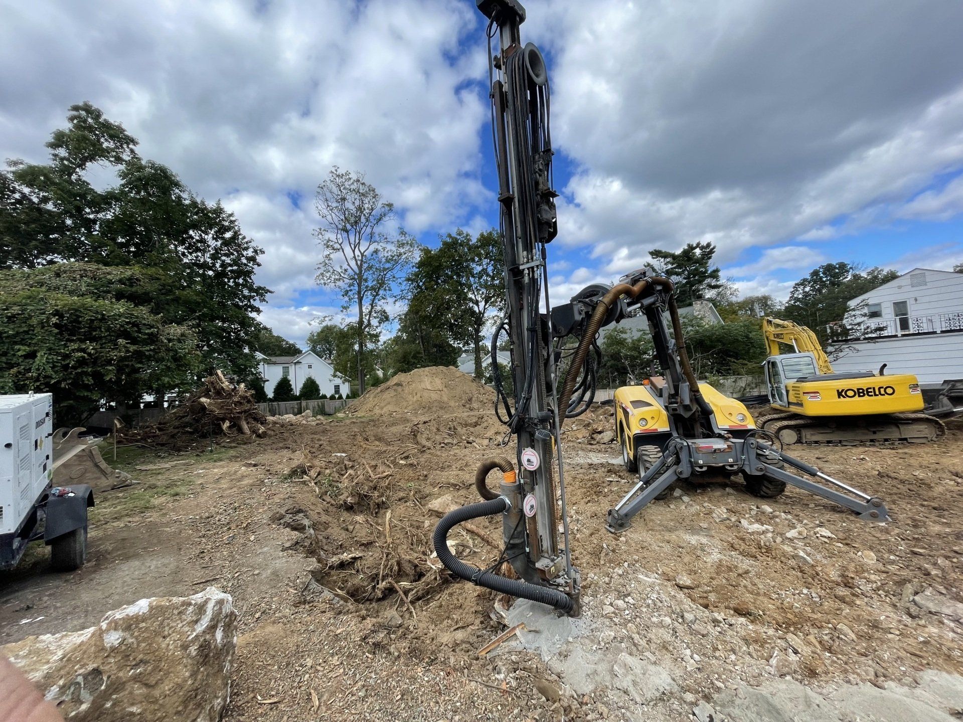 Construction site with drilling rig, excavator, and pile of dirt under cloudy sky.