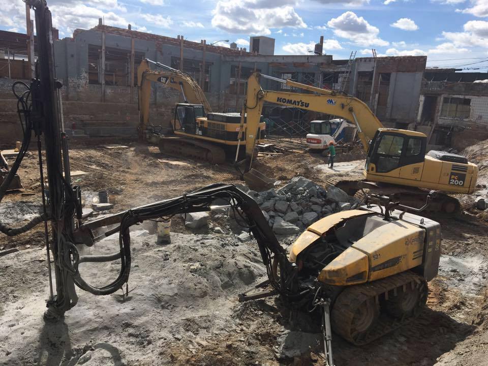 Construction site with multiple excavators and drilling equipment. Building in background.