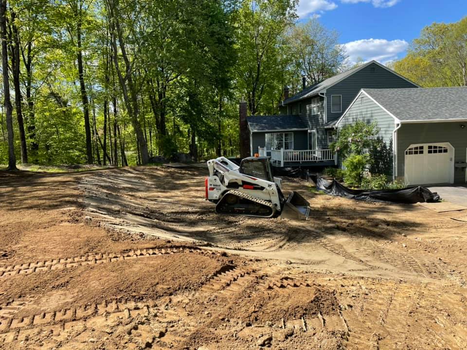 A small Bobcat machine on dirt clearing land near a two-story house with trees in the background.